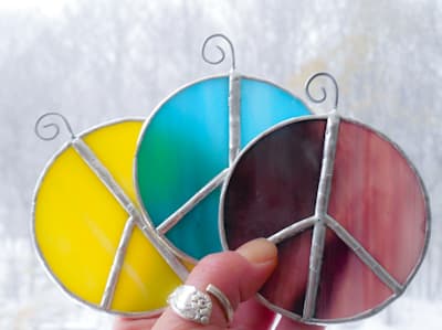 Hand holding three peace sign stained glass suncatchers in aqua, yellow and purple